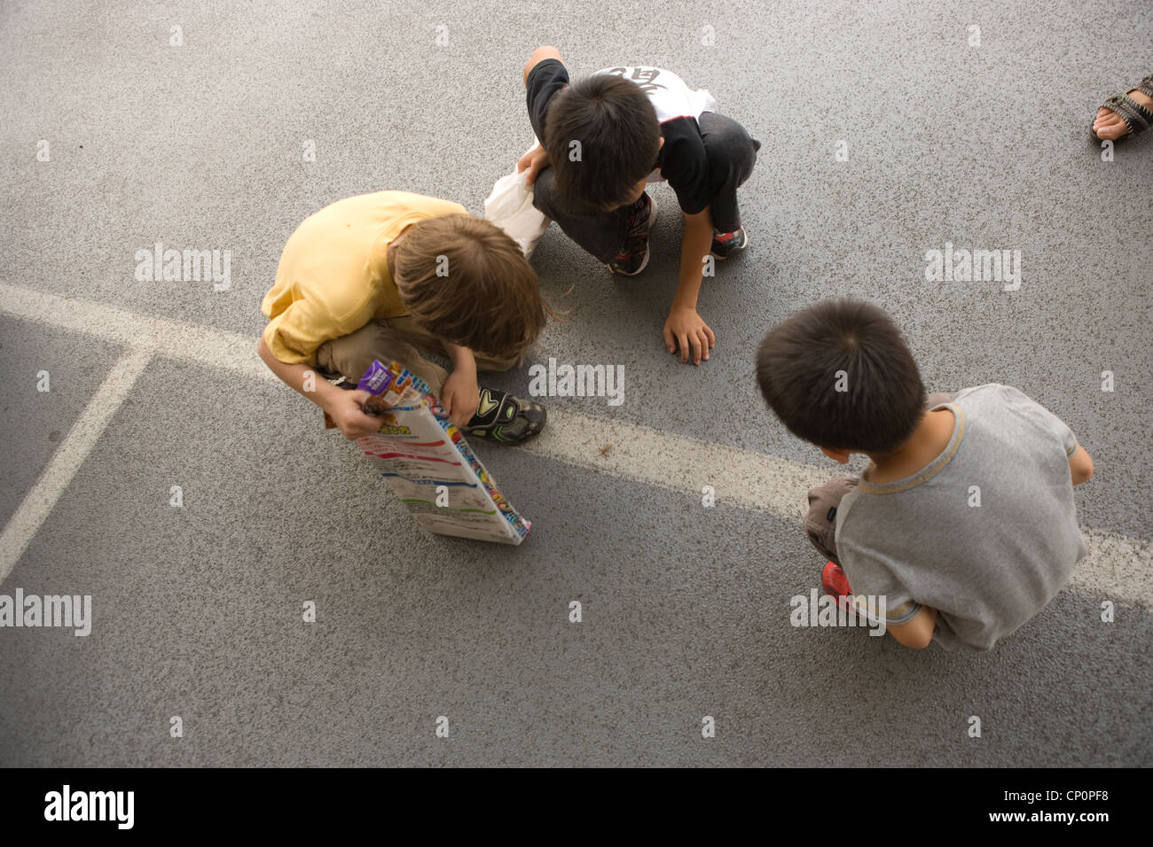 Three boys squat down in a parking lot to study an insect walking. Stock Photo