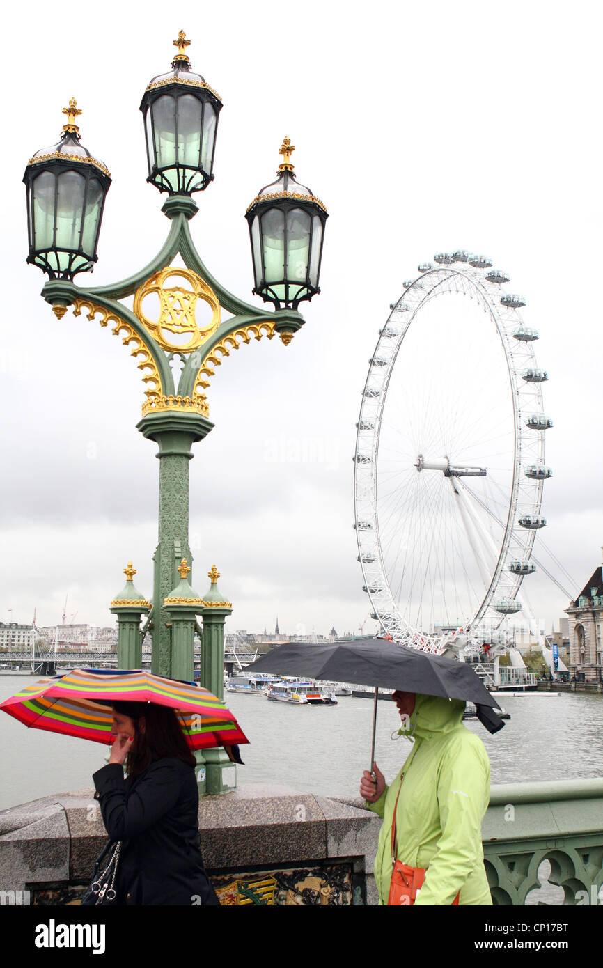 two women, with umbrellas, walking over Westminster Bridge with the London Eye in the background Stock Photo
