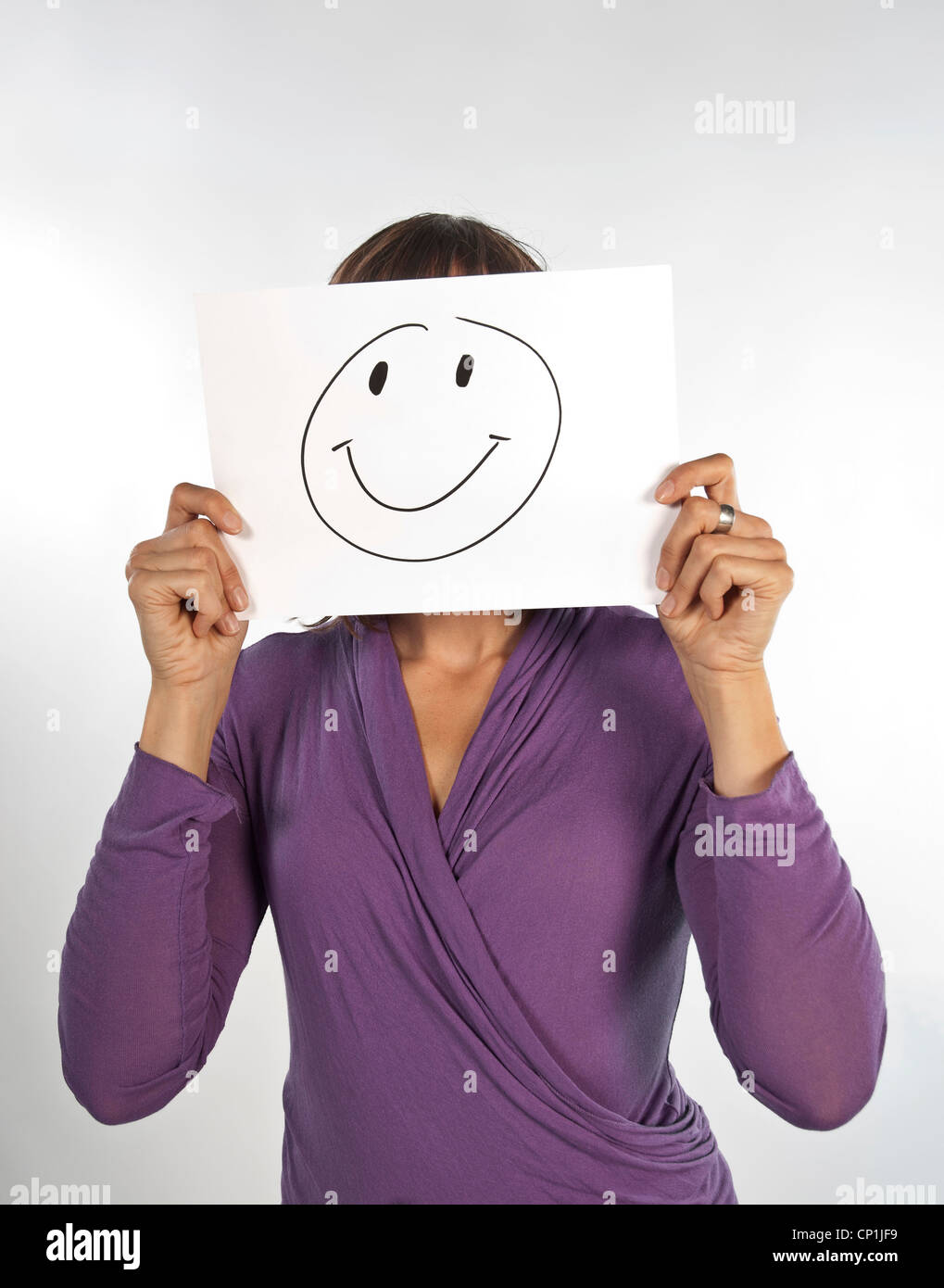 Woman hides her face behind a sheet of paper on which a smiley is drawn. Stock Photo