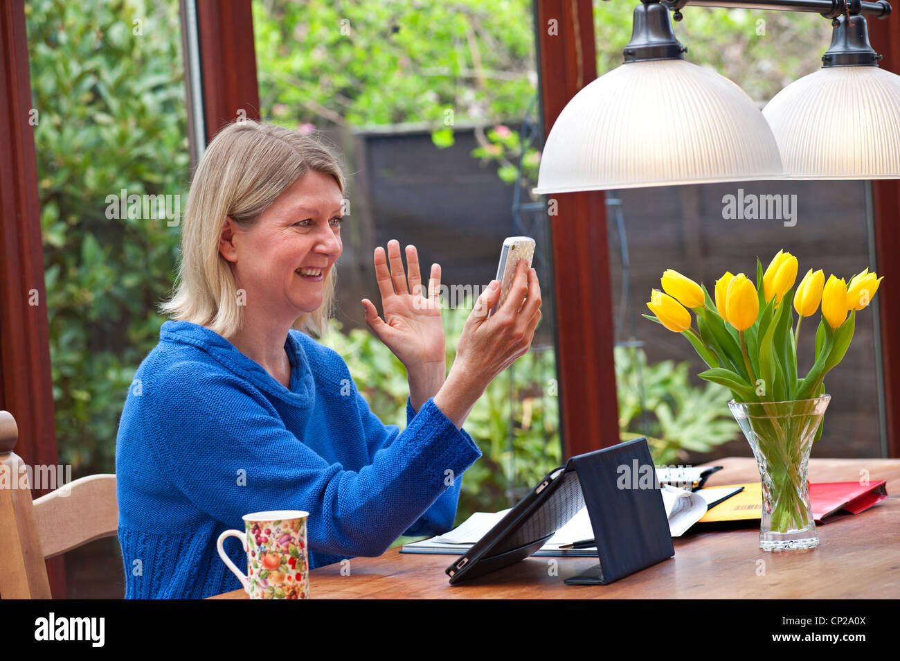 Blond woman at home in garden conservatory using her iPad tablet computer and using video call on her iPhone 4s smartphone Stock Photo