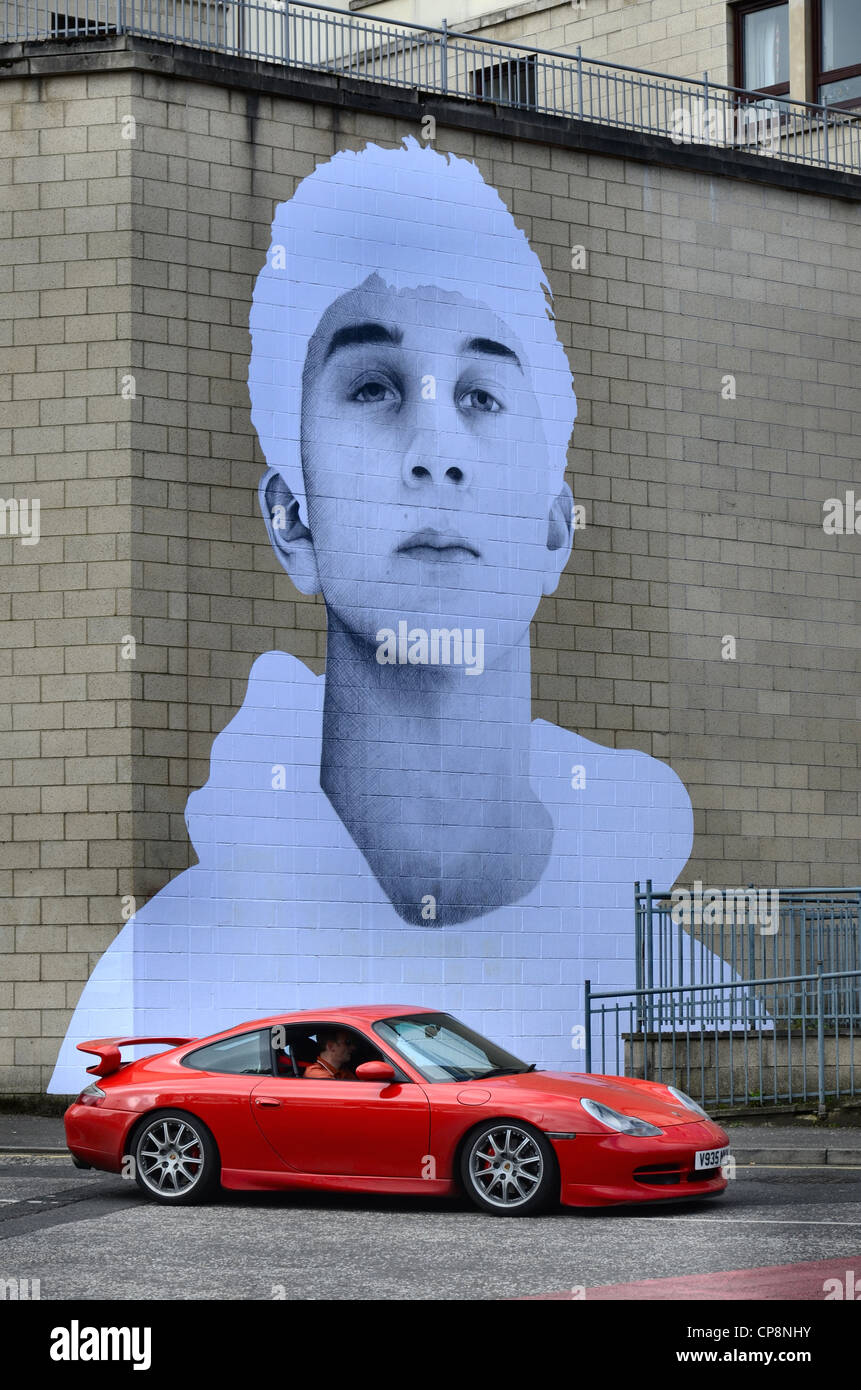 A red sportscar parked in front of a sticker depicting a teenage boy, part of a community based arts project. Stock Photo