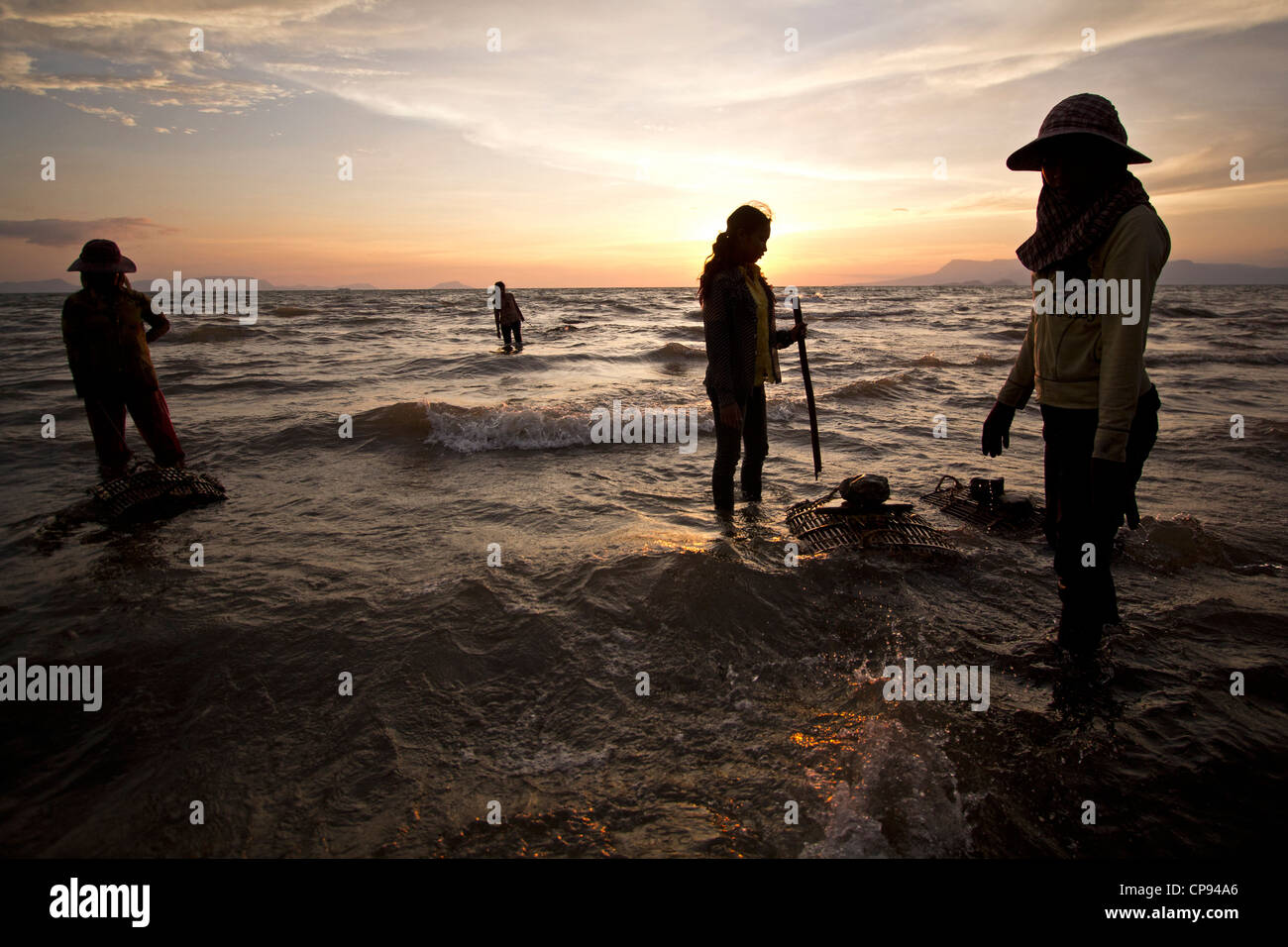 Female crab sellers stand in the water in Kep, Cambodia, with the sun setting behind them over the Gulf of Thailand Stock Photo