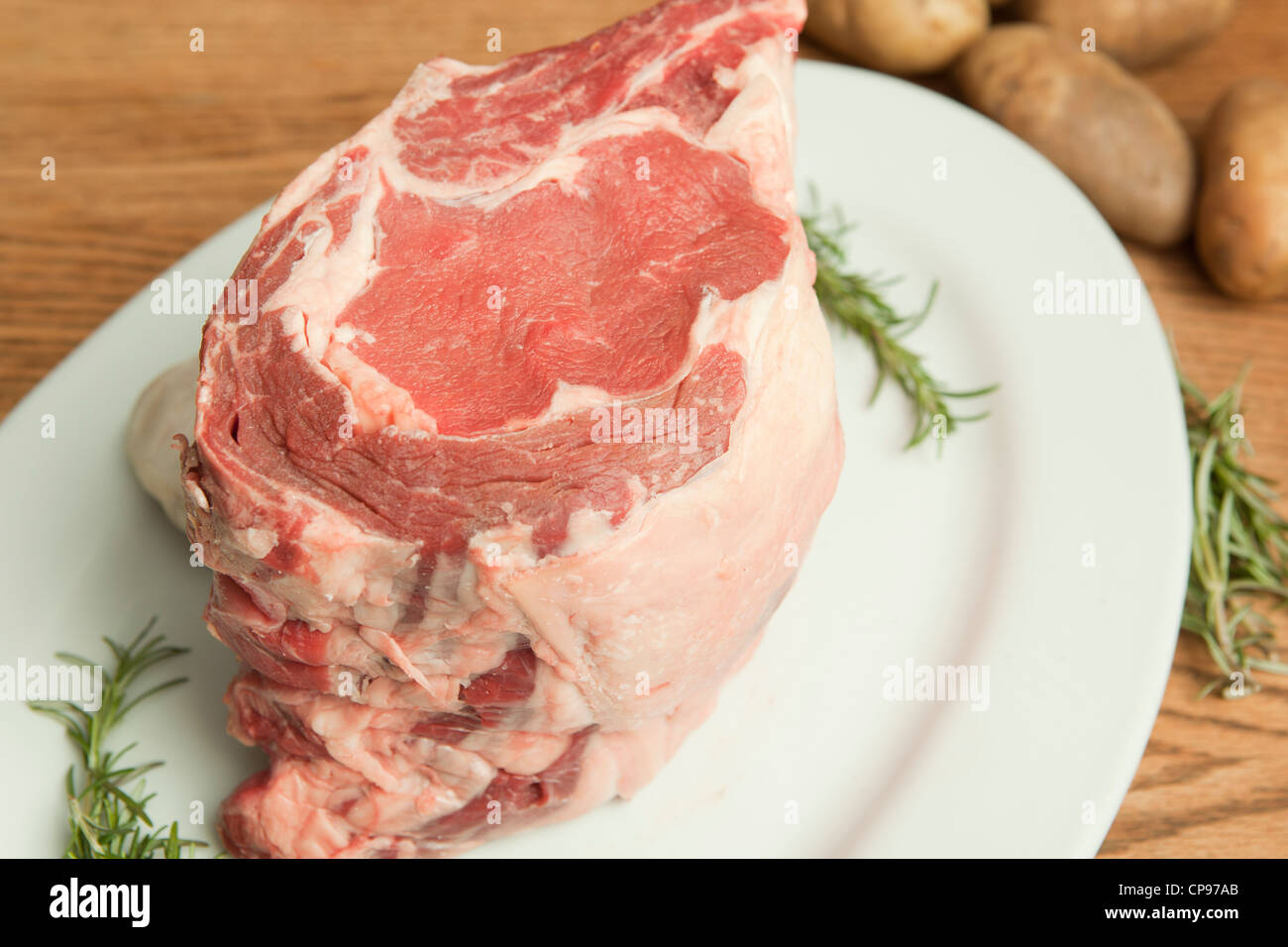 Horizontal view of a large uncooked prime rib roast along with garnishes of herbs, garlic and potatoes Stock Photo