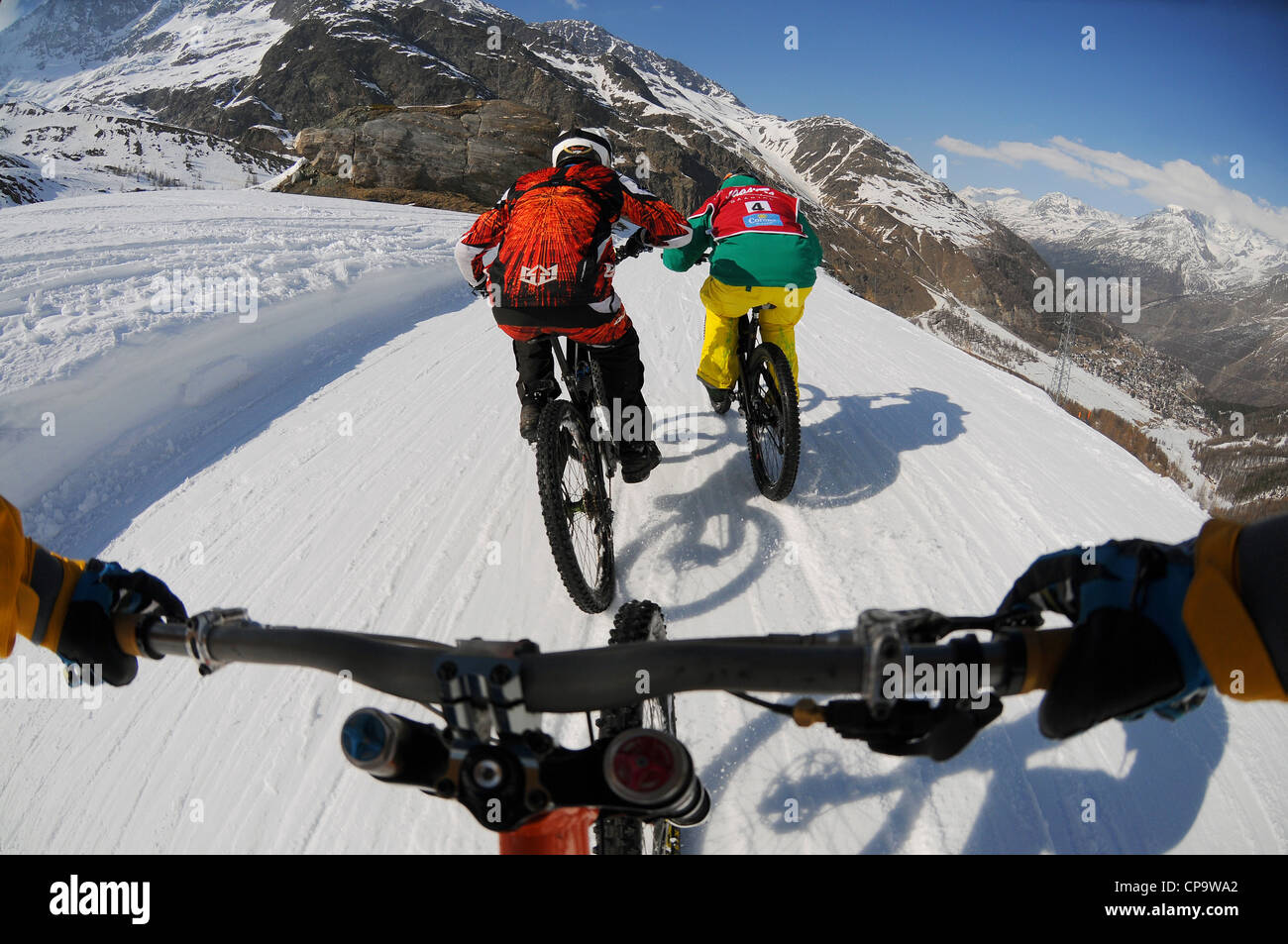 A mountain biker rides downhill hill on snow with an on-board camera following two other riders. Stock Photo