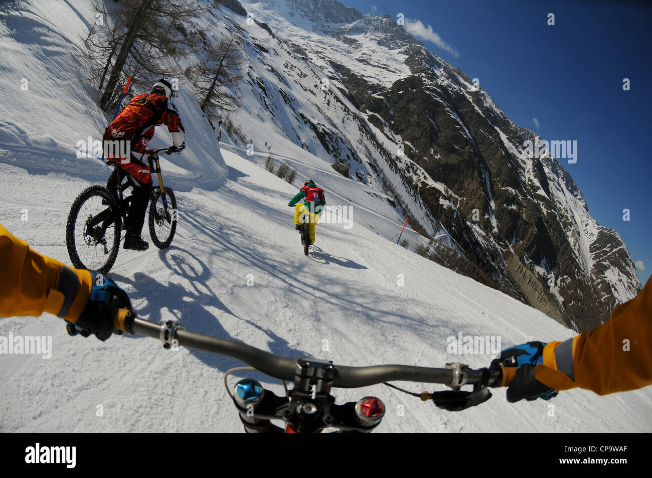 A mountain biker rides downhill hill on snow with an on-board camera following two other riders. Stock Photo