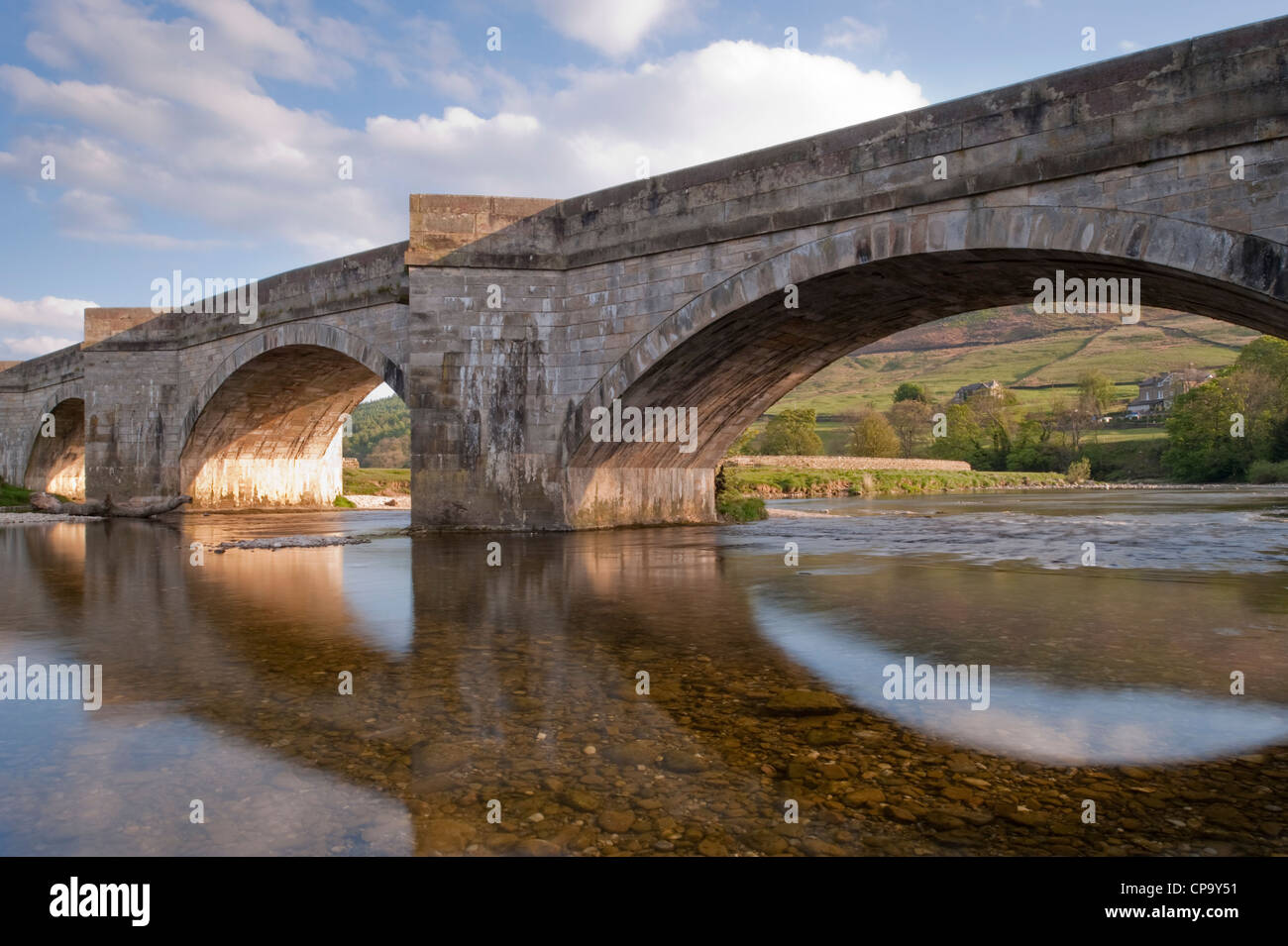 Sunny evening, riverside view of old stone, arched bridge reflected in  shallow, clear water of River Wharfe, Burnsall, Yorkshire Dales, England, UK. Stock Photo