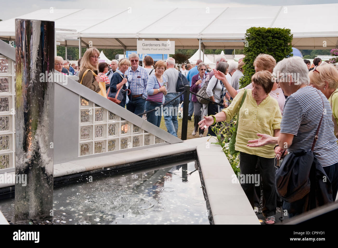 Crowd of people viewing SHINE Garden for Cancer Research UK, as woman throws coin into water pond - RHS Flower Show, Tatton Park, Cheshire, England. Stock Photo