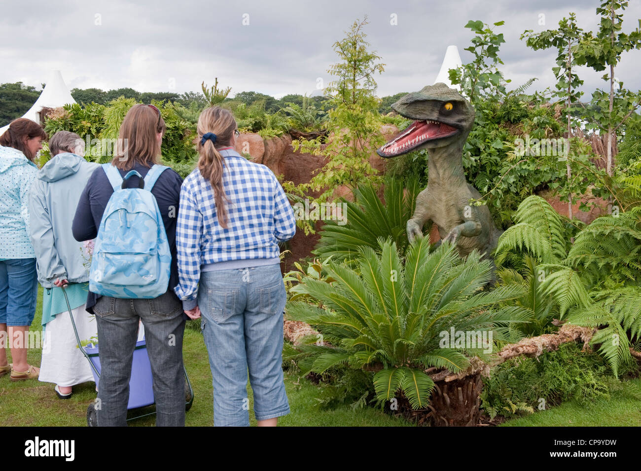 People view fierce raptor, green ferns & palms in prize-winning show garden (Dinosaurs at Large exhibit) - RHS Flower Show, Tatton Park, Cheshire, UK. Stock Photo