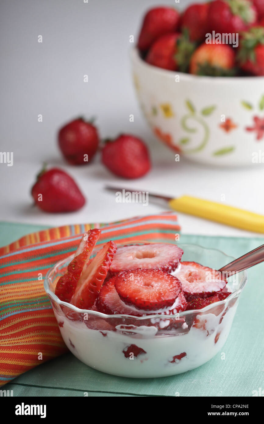 Glass bowl of fresh sliced strawberries with cream and sugar, decorative bowl of whole strawberries in the background. Stock Photo