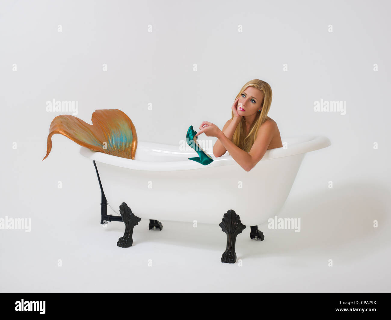 Young blond mermaid sitting in her  bathtub holding up a teal high heel shoe looking longingly off into the distance Stock Photo