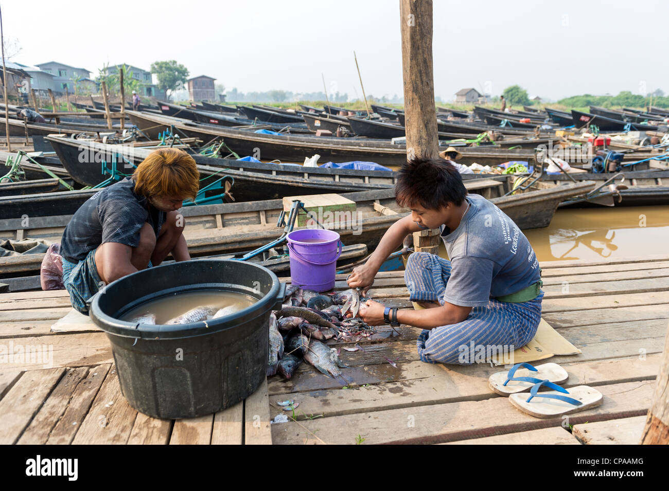 Two young men cleaning and preparing fish on a wooden dock beside fishing boats in Phaung Daw Oo pagoda marketplace in Shan, Myanmar.Burma Stock Photo