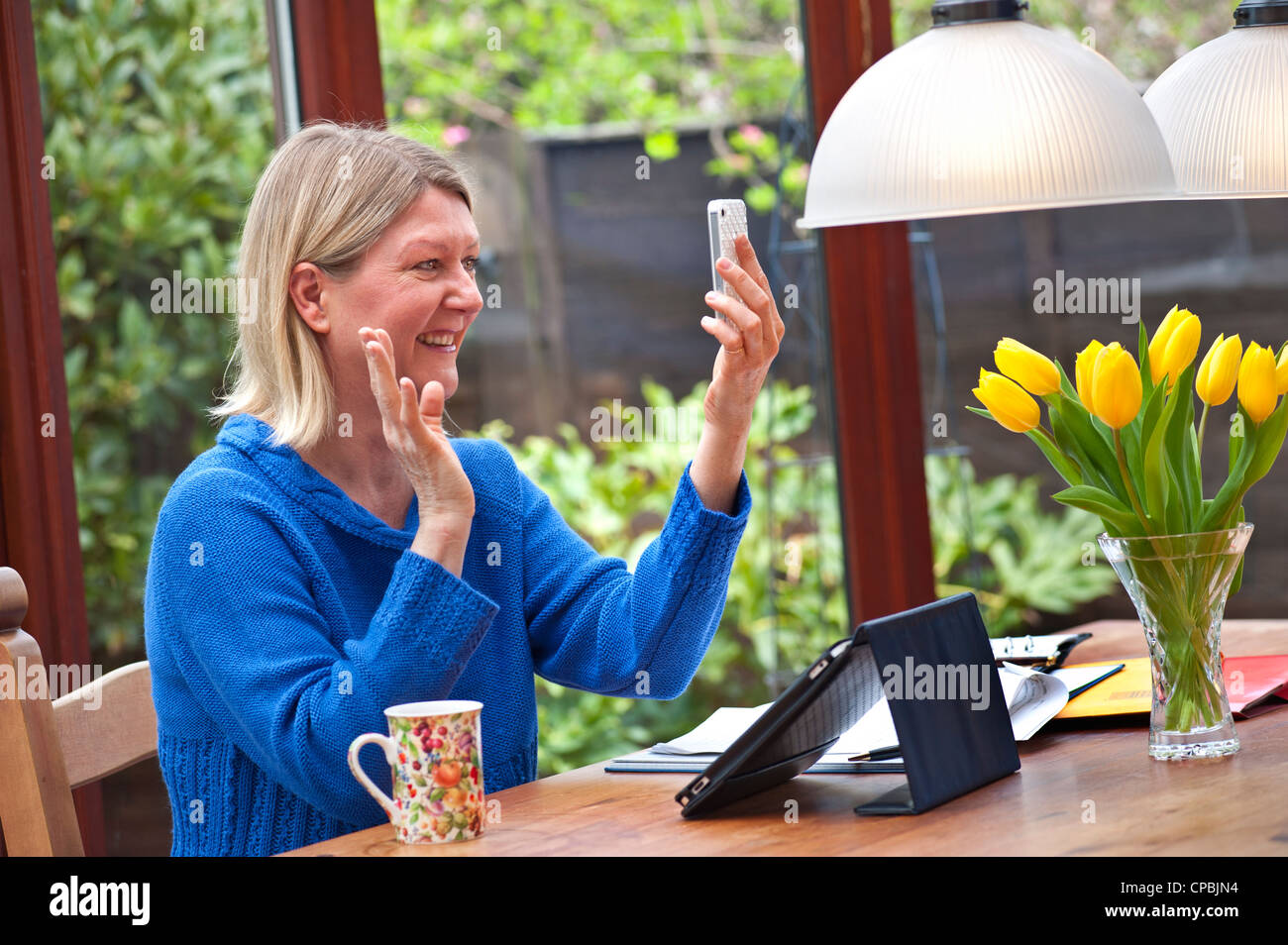 FaceTime Zoom video call Blond woman at home in spring garden conservatory using her iPad tablet computer and using video call on iPhone smartphone Stock Photo