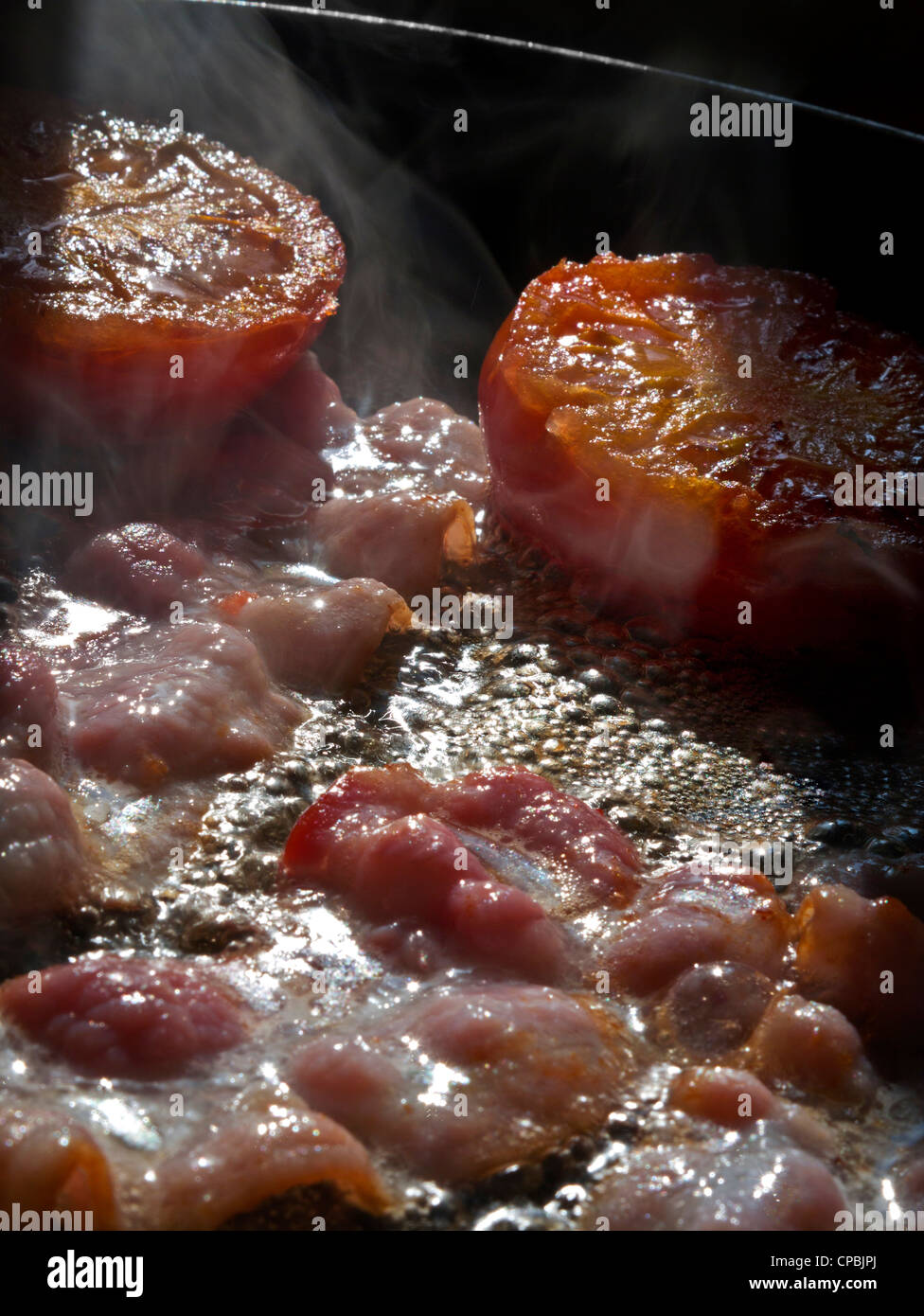 Shaft of sunlight illuminates streaky bacon rashers and tomatoes frying in hot pan Stock Photo