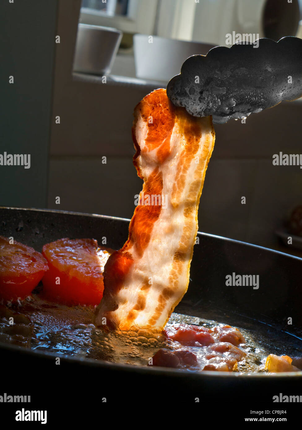 STREAKY BACON FRYING PAN KITCHEN in sunlight illuminating a  rasher of organic streaky bacon being placed into a hot frying pan containing tomatoes Stock Photo