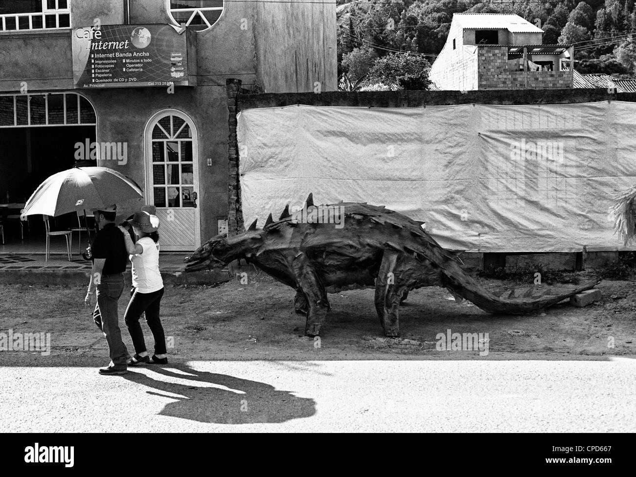 Couple in the street with an umbrella walking by a dinosaur's model. Sutamarchan, Boyacá, Colombia, South America, Andes Stock Photo