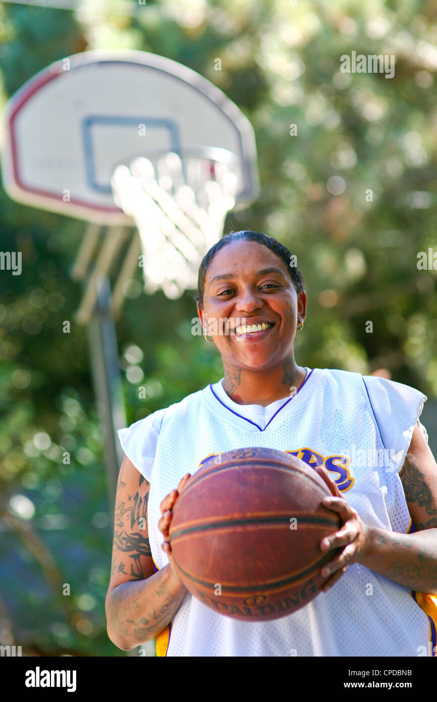 A young African-American woman with tattoos smiles, holding a basketball in front of a basketball hoop. Stock Photo