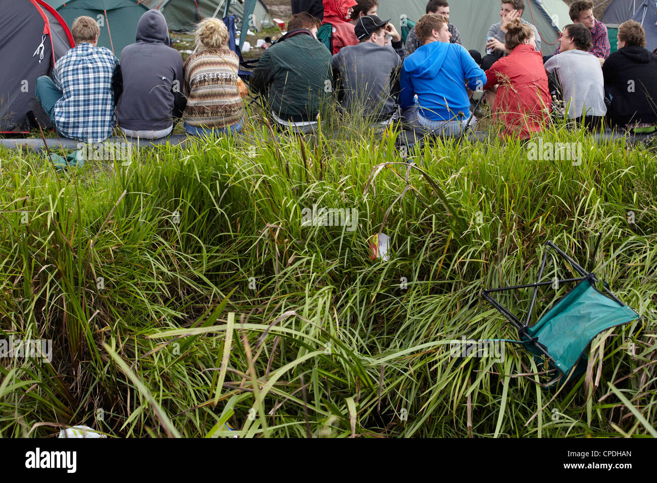 Row of festival goers sitting and talking with backs facing. A broken chair tumbles down the ditch in the foreground. Stock Photo