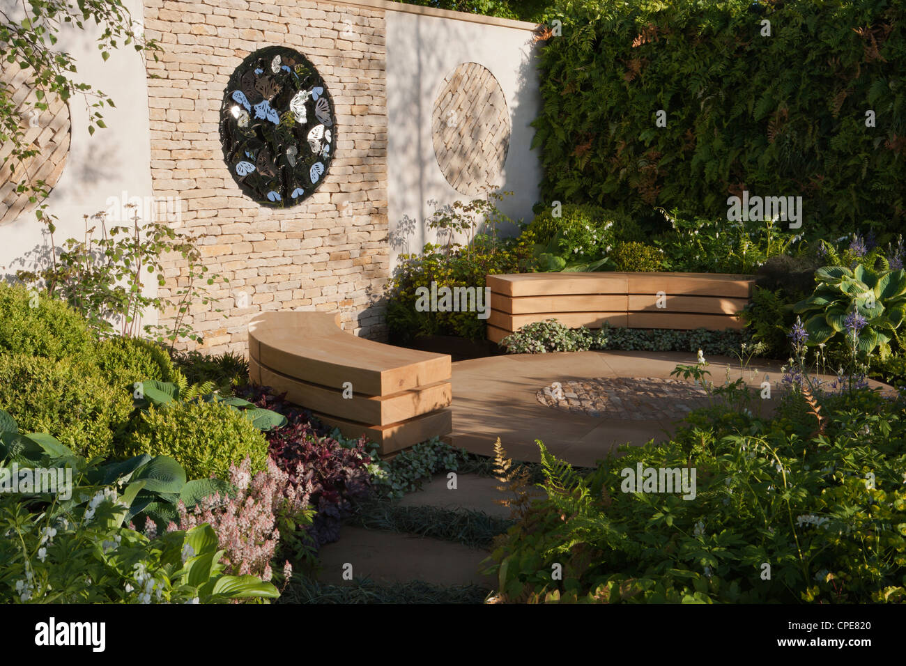 Cottage courtyard garden with cotswold dry stone wall and circular circle patio area and wooden wood bench benches and a hedge boundary UK Stock Photo