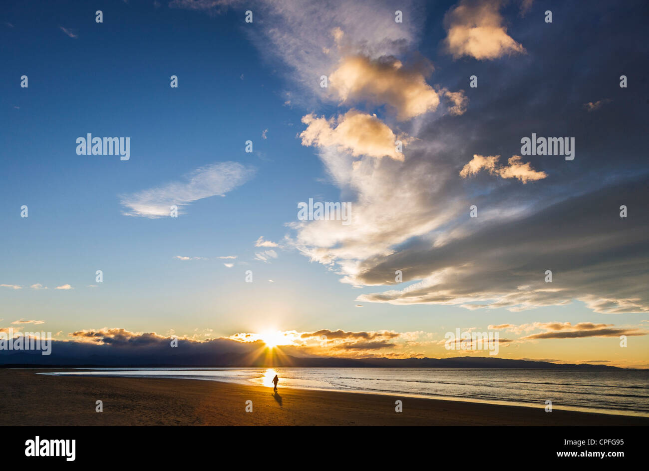 Tahuna Beach, Nelson Bay, New Zealand. Person walking on beach at edge of sea just as sun disappears behind mountains. Stock Photo