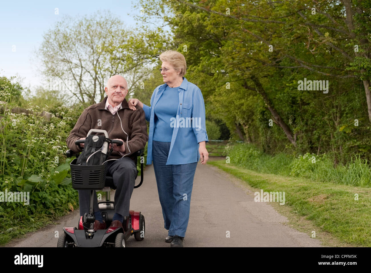 Mature happily married couple out for a stroll in the countryside while one drives a motorised mobility scooter Stock Photo