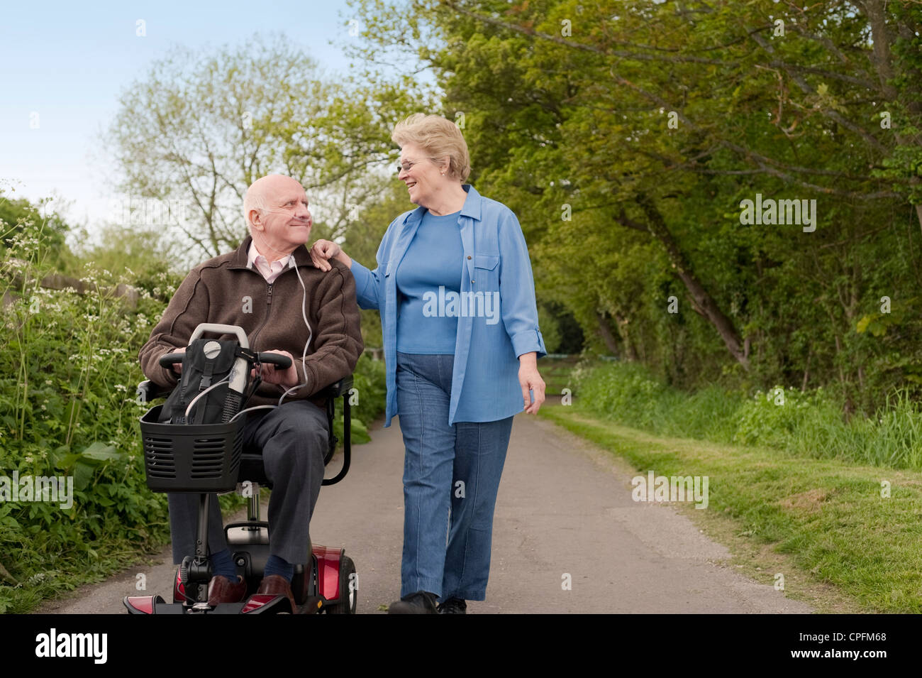 Mature happily married couple out for a stroll in the countryside while one drives a motorised electric mobility scooter Stock Photo