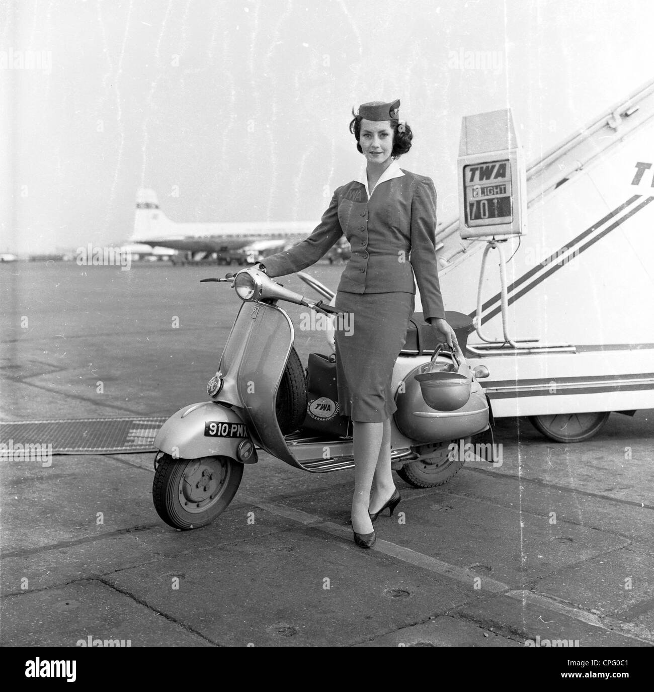 1960s, historical, an advertising still from this era promoting the new motor scooter, showing a glamorous TWA air stewardess at an airport, at the boarding steps of a plane, standing with helmet, by her new mode of transport, London Aiport, England, UK.. Stock Photo
