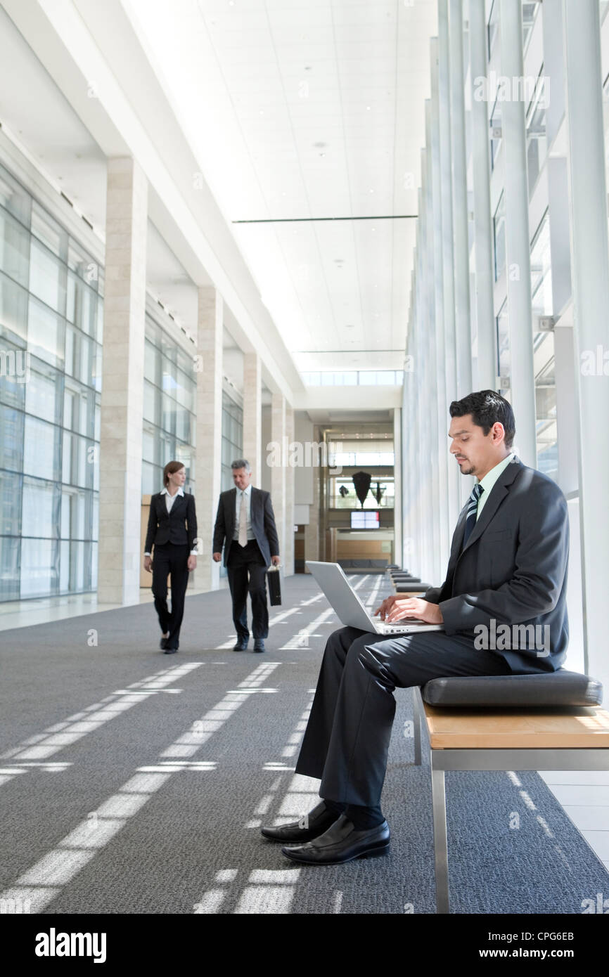 Businessman with laptop sitting on bench in office hallway, two business people talking by outdoor. Stock Photo