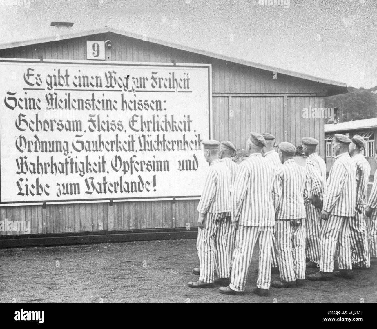 Camp internees in striped uniforms at Sachsenhausen concentration camp standing before a sign with a National Socialist slogan, Stock Photo