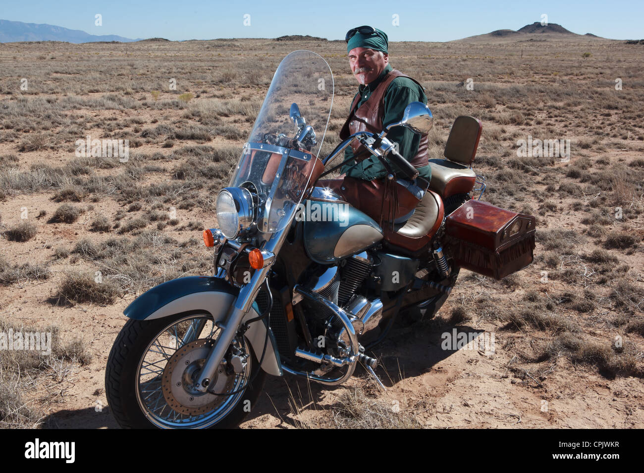 Older Caucasian man wearing brown leather leaning up against his motorcycle out in the desert outside of Albuquerque, New Mexico Stock Photo