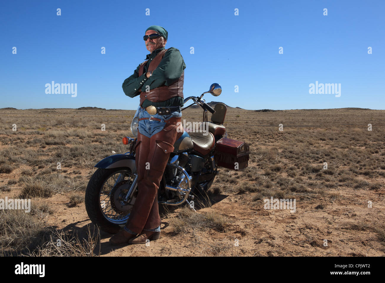 Older Caucasian man wearing brown leather standing next to his motorcycle out in the desert. Stock Photo