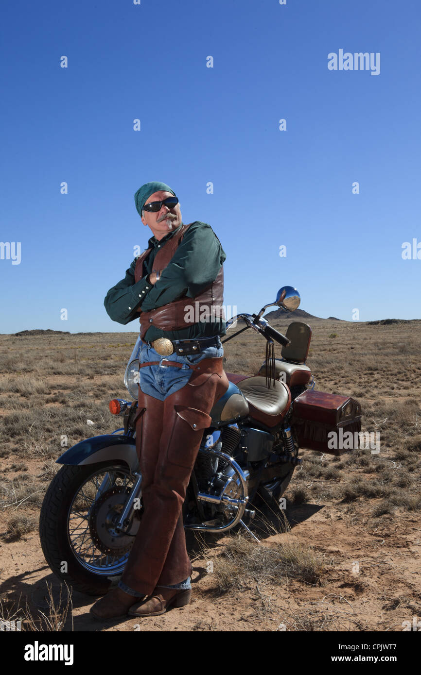 Older Caucasian man wearing brown leather standing next to his motorcycle out in the desert. Stock Photo