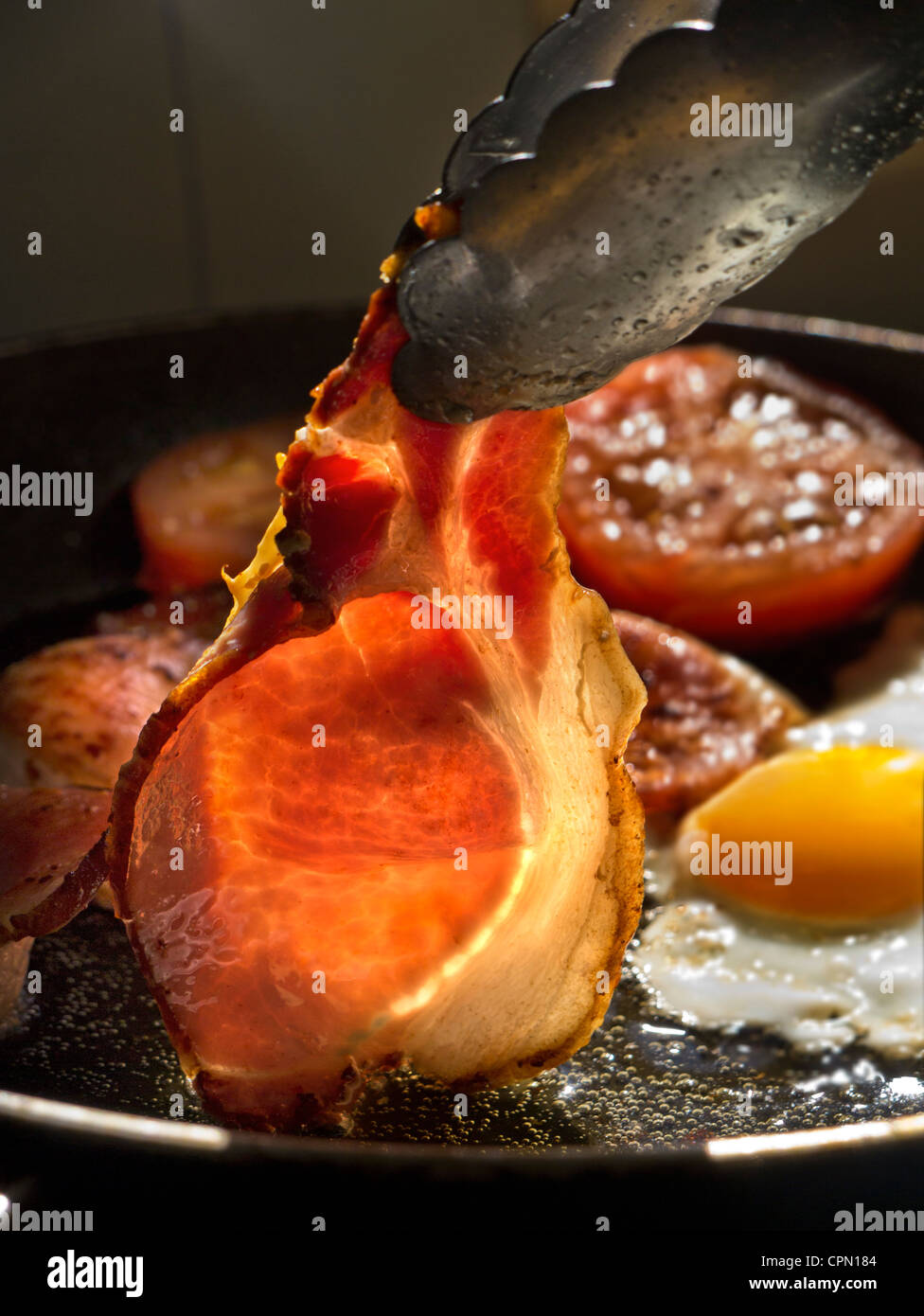 BACON EGGS TOMATOES FRYING in sunlight illuminating a rasher of organic back bacon being turned in a hot frying pan containing fried egg and tomatoes Stock Photo