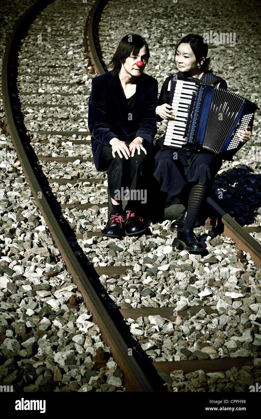 a clown couple sitting on railroad tracks, the woman plays music Stock Photo
