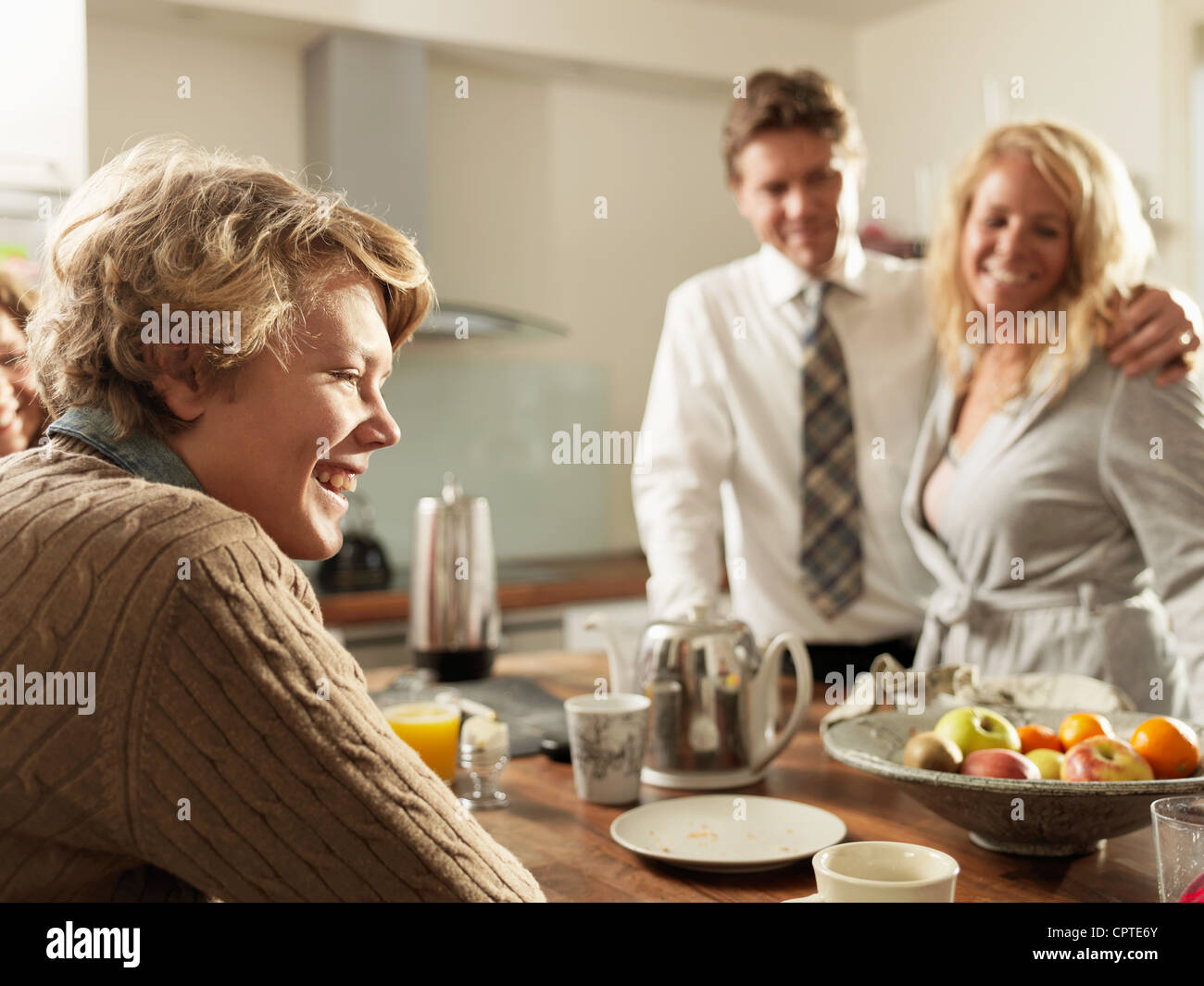 Teenage son sitting at kitchen table with parents in background Stock Photo