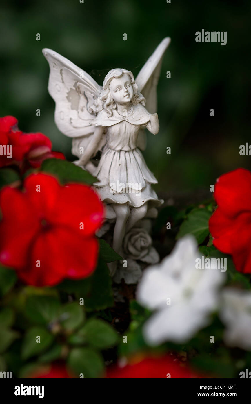 A small, stone angel or fairy figurine with wings among red and white flowers. Stock Photo