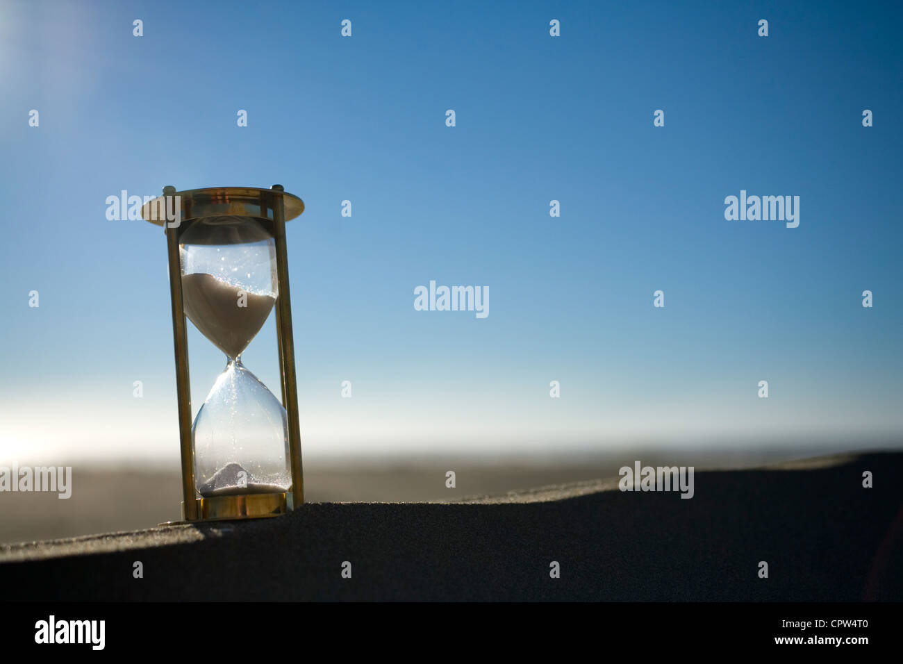 Backlit sand timer on the peak of a sand dune, with sand and sky behind it. Stock Photo