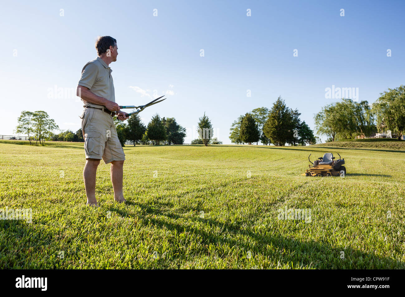 Challenging task of cutting large lawn with grass shears by hand with mower in background Stock Photo