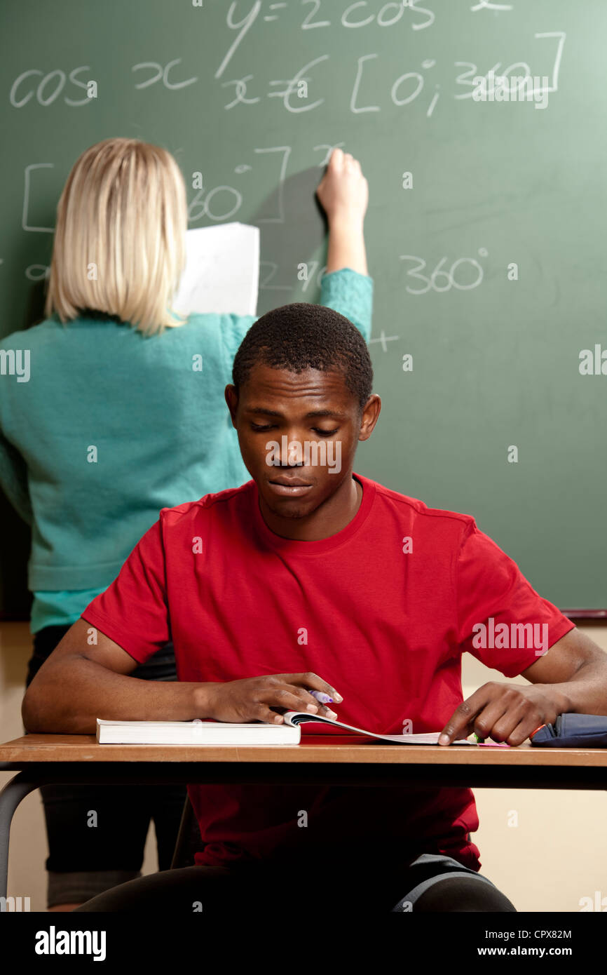 Male student working at a desk with teacher in background. Stock Photo