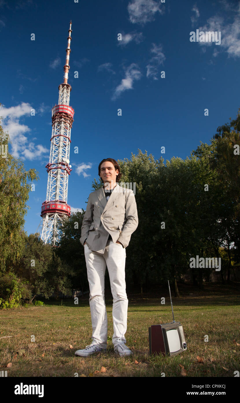 Young man with portable retro tv set in front of high television tower holding his hands in pockets Stock Photo