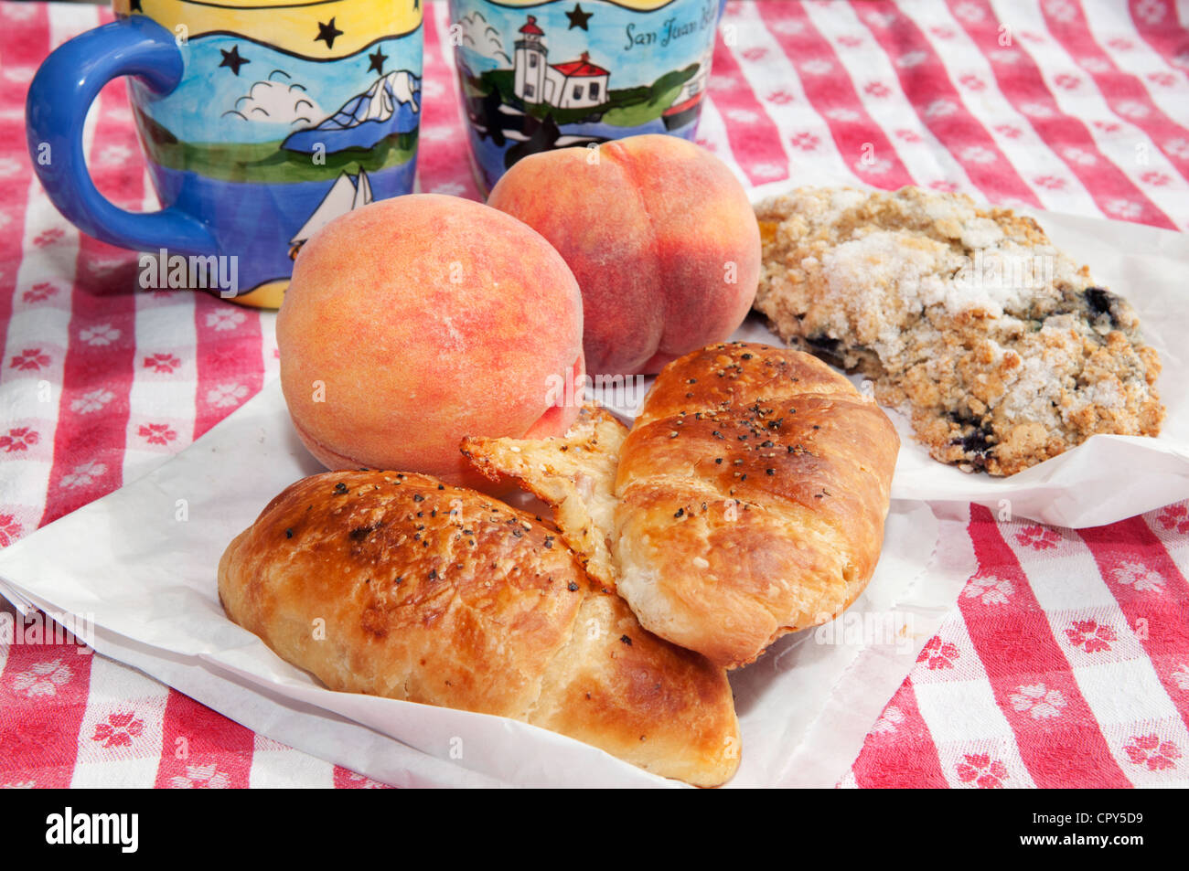 Picnic breakfast of fresh peaches, croissants, a scone and coffee is served on a checkered tablecloth. Stock Photo