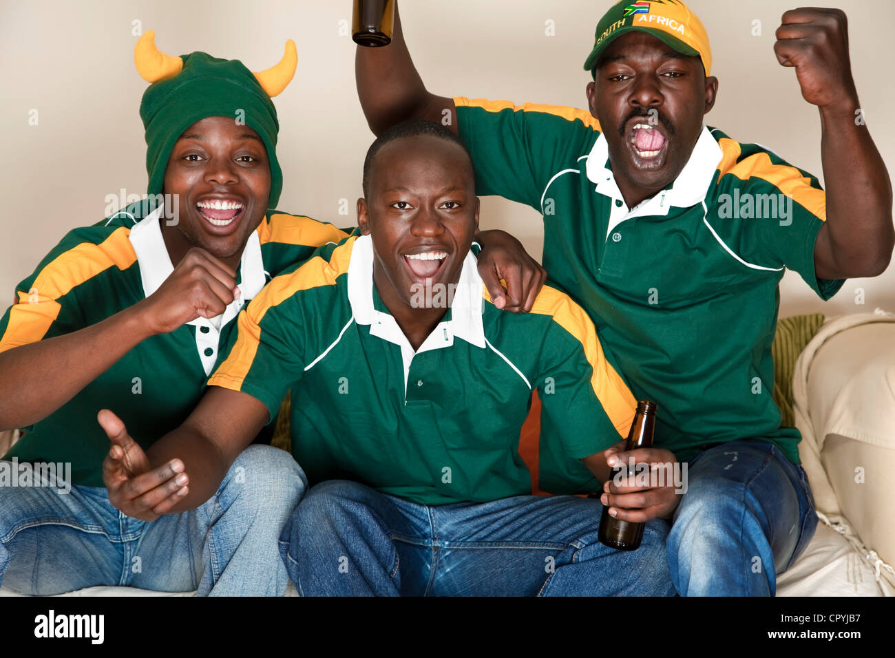 Three friends dressed in rugby uniforms watch a rugby game on TV whilst ...