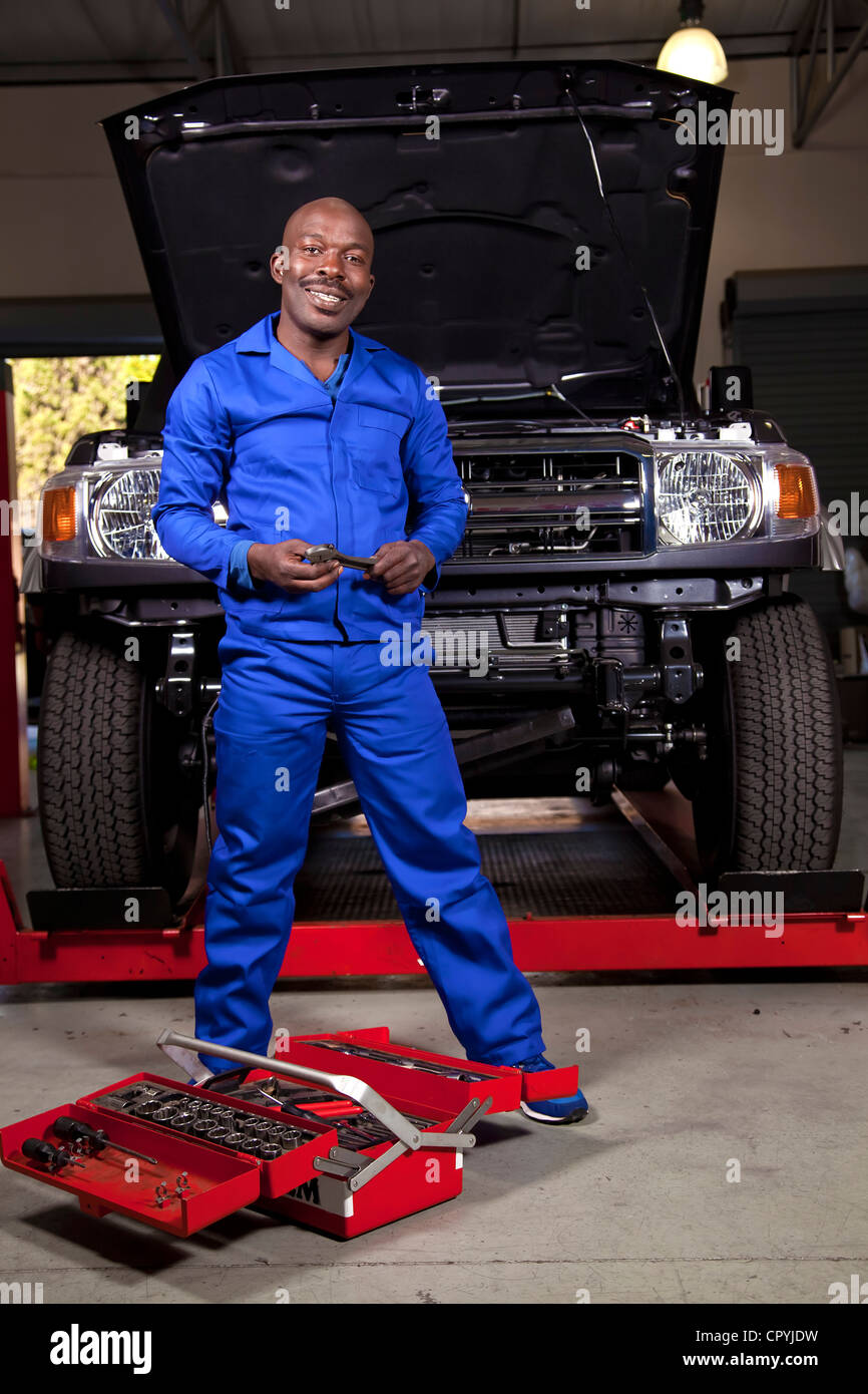An African mechanic standing in front of a car with a toolbox, smiling