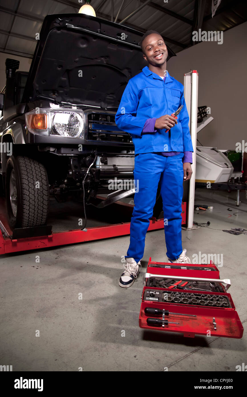 An African mechanic standing in front of a car with a toolbox, smiling at camera Stock Photo
