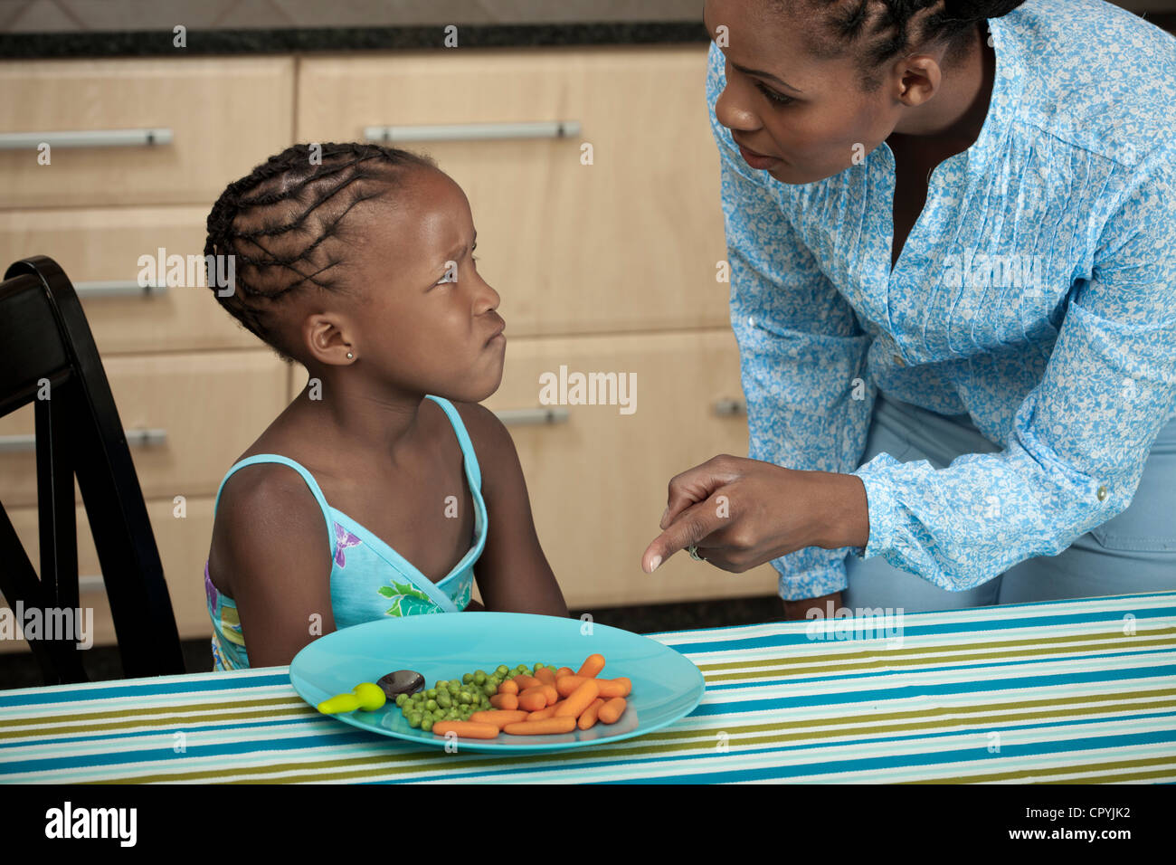 Young African girl frowning while her mother tells her to eat her vegetables Stock Photo
