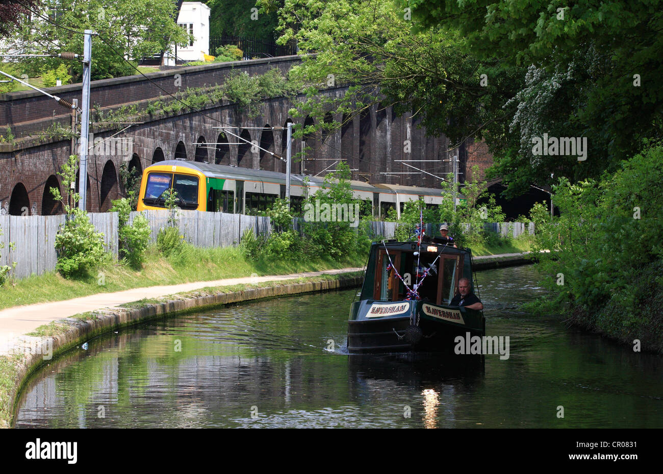 a canal boat traveling along a canal in Birmingham with a train traveling in the background Stock Photo