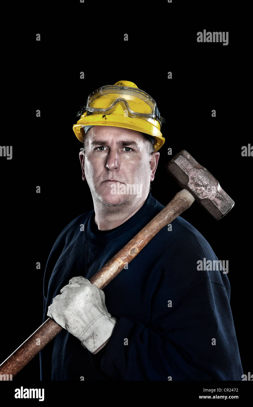 A manual laborer with a sledgehammer wearing a yellow hardhat and protective work gloves isolated on black Stock Photo