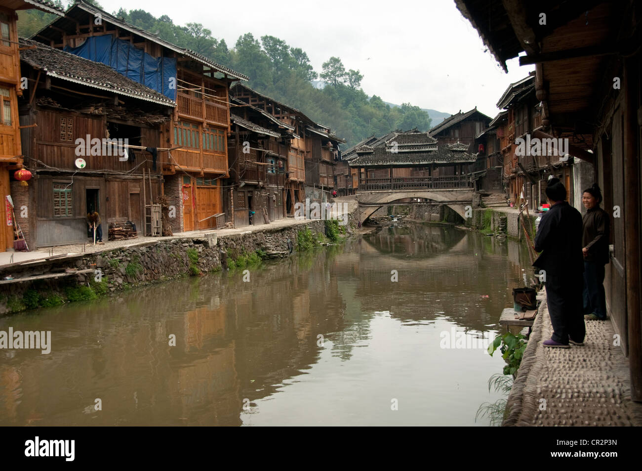 Two Dong women talking on a narrow lane on the side of a stream, a small 'Wind and Rain' bridge, Zhaoxing Dong Village, China Stock Photo