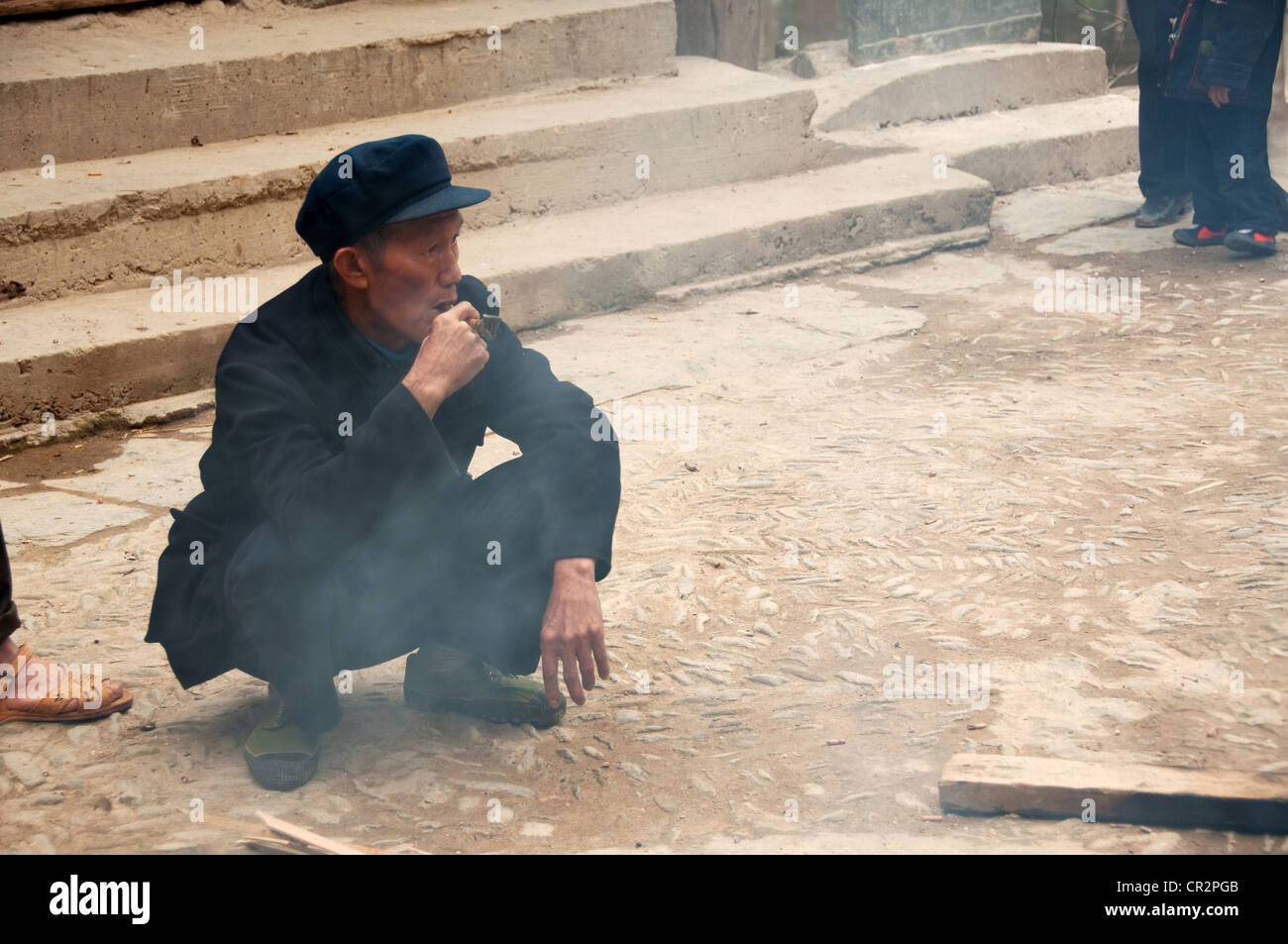 A mature man squat with a hat smoking a pipe, Zhaoxing Dong Village, Southern China Stock Photo