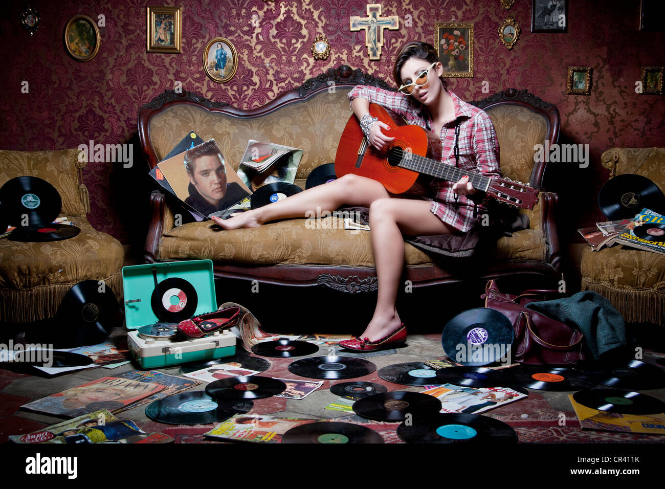 Girl playing guitar on an old sofa, with many vinyls and magazines on the floor. A retro style shot, perfect for events adverts. Stock Photo