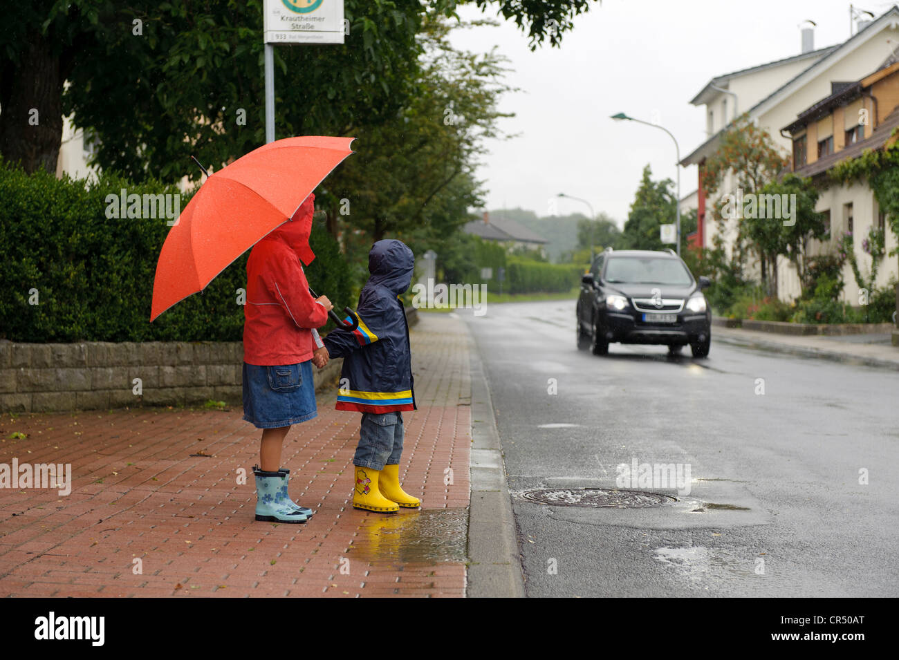 Two children, 3 and 7 years, waiting to cross the street in the rain while a car approaches, Assamstadt, Baden-Wuerttemberg Stock Photo